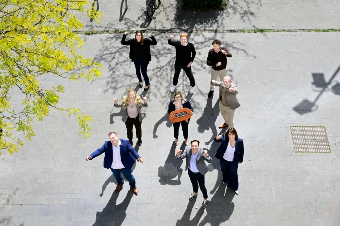 Nine people stand on pavement, looking up with raised arms